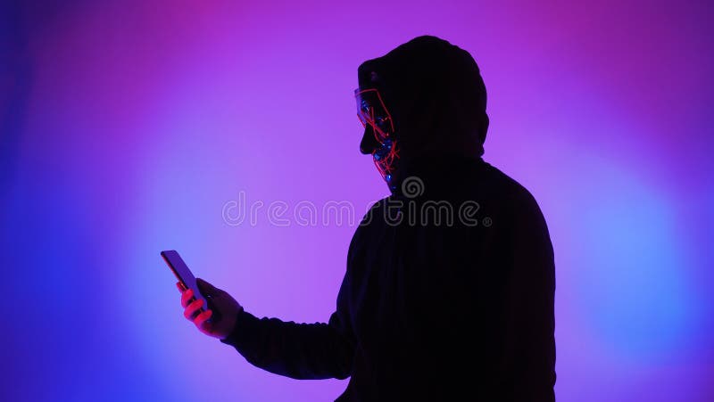 Anonymous hacker and face mask with smartphone in hand. Man in black hood shirt trying to hack personal data from mobile phone. Represent cyber crime data hacking or personal data stealing concept. Anonymous hacker and face mask with smartphone in hand. Man in black hood shirt trying to hack personal data from mobile phone. Represent cyber crime data hacking or personal data stealing concept