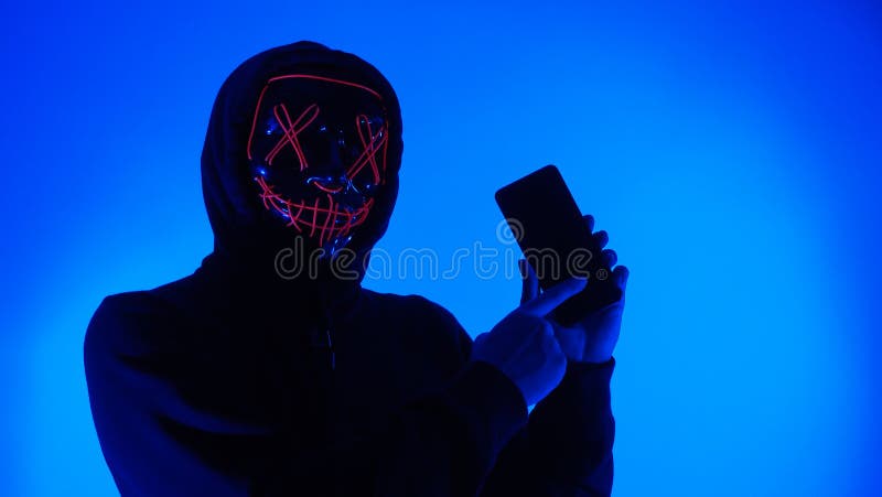 Anonymous hacker and face mask with smartphone in hand. Man in black hood shirt trying to hack personal data from mobile phone. Represent cyber crime data hacking or personal data stealing concept. Anonymous hacker and face mask with smartphone in hand. Man in black hood shirt trying to hack personal data from mobile phone. Represent cyber crime data hacking or personal data stealing concept