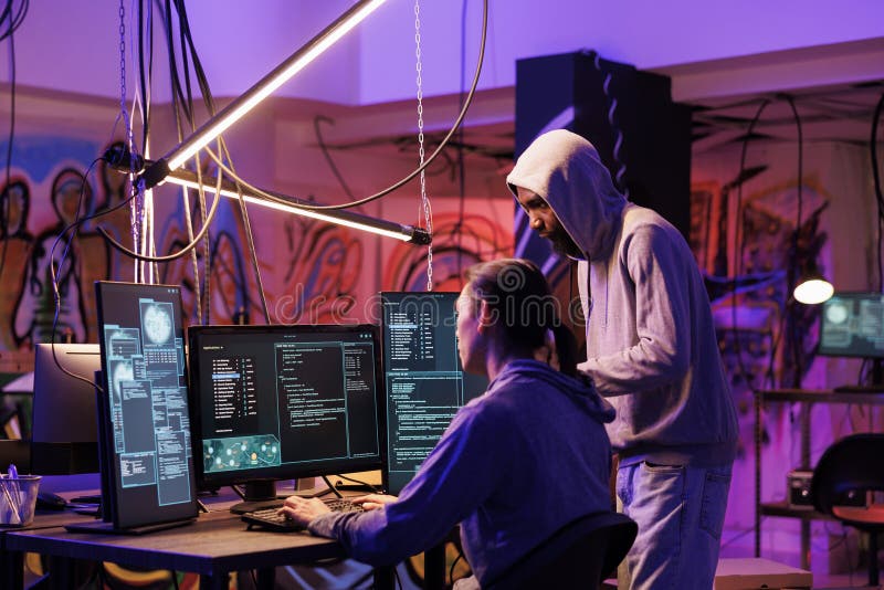 Hackers coding malicious software on computer together while working in abandoned warehouse. Internet thieves in hoods planning phishing attack and programming malware in dark room