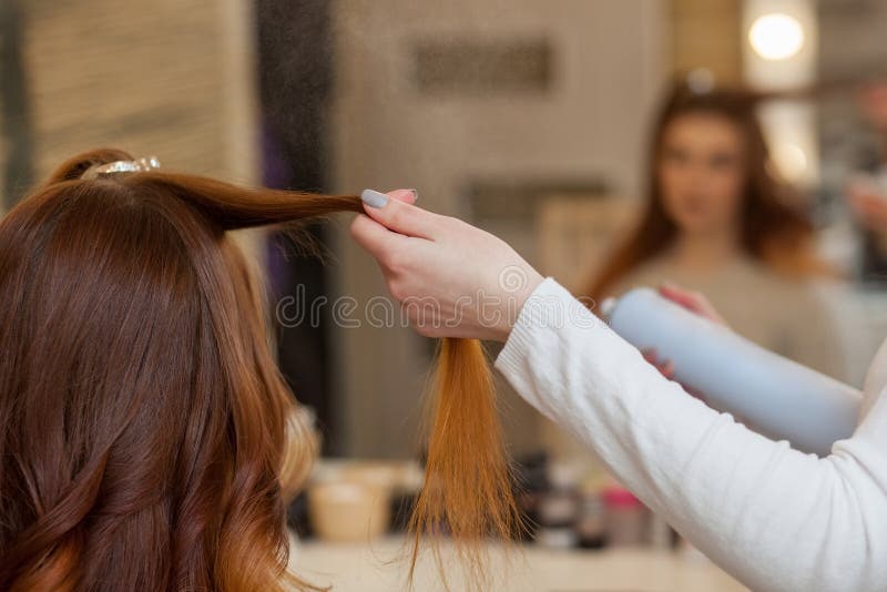 Hairdresser combing her long, red hair of his client and sprinkles hairspray in a beauty salon.