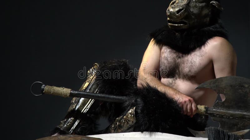 The hairy man in the mask of the minotaur sits with an ax in his hands around the scattered arms and armor