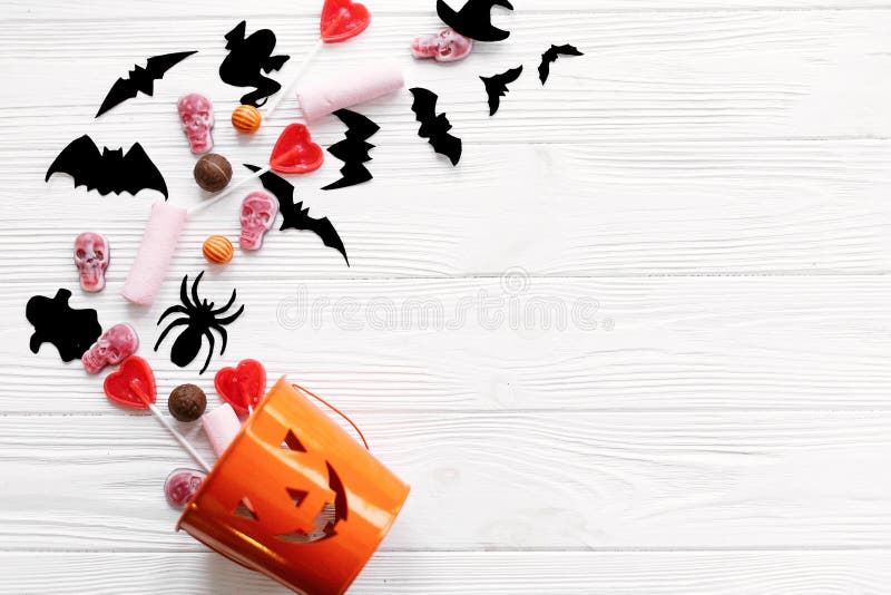 Halloween candy with skulls, black bats, ghost, spider decorations spilled from jack o lantern bucket on white wooden background, flat lay. Halloween sweets. Copy space. Trick or treat. Halloween candy with skulls, black bats, ghost, spider decorations spilled from jack o lantern bucket on white wooden background, flat lay. Halloween sweets. Copy space. Trick or treat