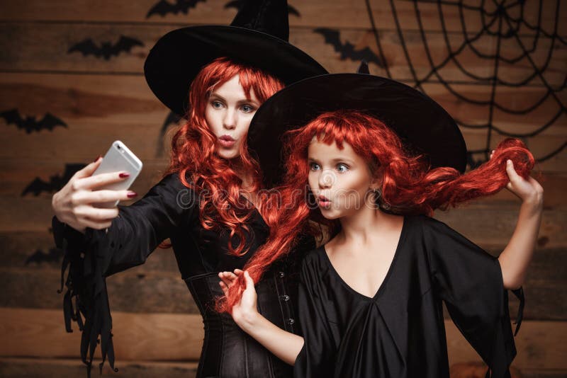 Halloween-Konzept - schöne kaukasische Mutter und ihre Tochter mit dem langen roten Haar in den Hexenkostümen, die ein selfie neh lizenzfreie stockfotografie Halloween-Konzept - schöne kaukasische Mutter und ihre Tochter mit dem langen roten Haar in den Hexenkostümen, die ein selfie neh lizenzfreie stockfotografie