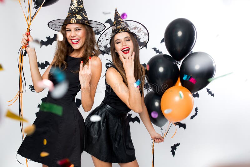 Halloween party. Two gorgeous girls in black dresses and witch hats have fun with balloons and confetti. .