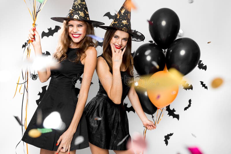 Halloween party. Two gorgeous girls in black dresses and witch hats have fun with balloons and confetti. .