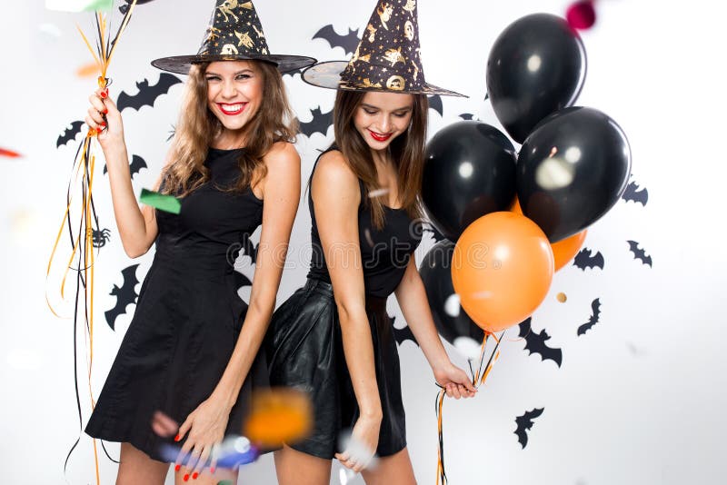 Halloween party. Two gorgeous girls in black dresses and witch hats have fun with balloons and confetti. .