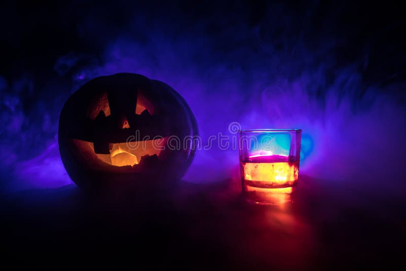 Halloween pumpkin with carved face and glass of whiskey with ice on a dark toned foggy background with zombies. Decorated.