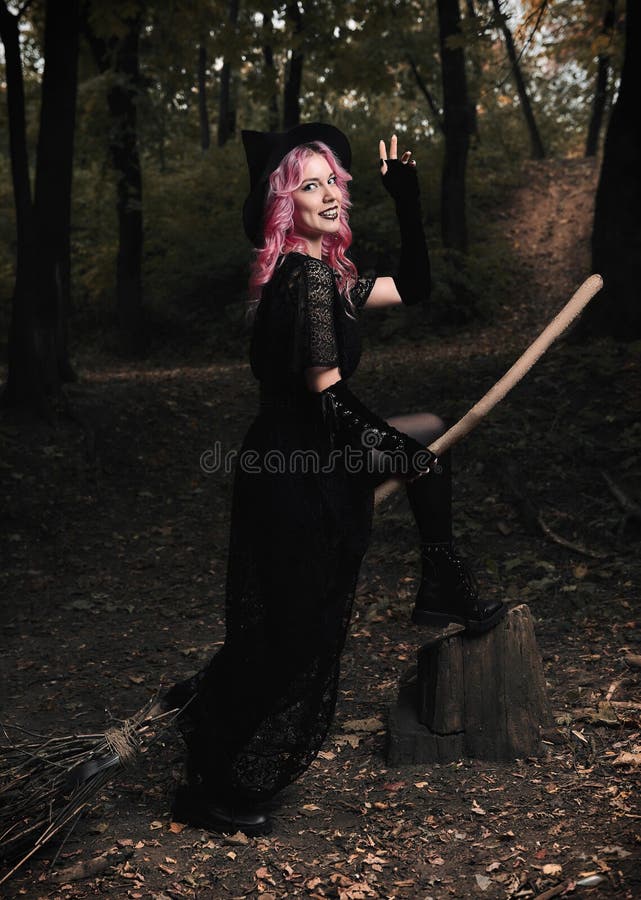 Halloween theme: modern smiling young witch in black dress and hat with broom in dark forest stock images Halloween theme: modern smiling young witch in black dress and hat with broom in dark forest stock images