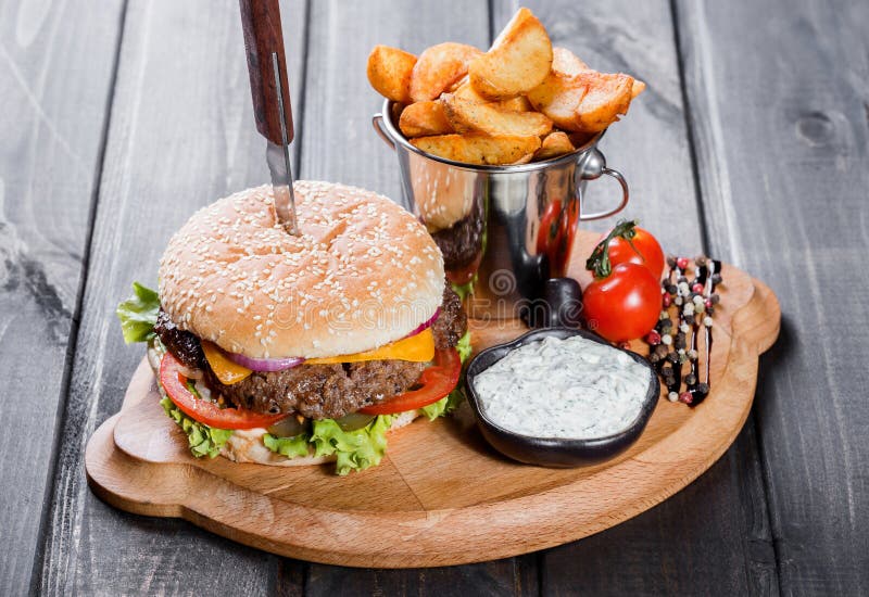 Hamburger with fresh vegetables, cheese, sauce and fries on cutting board on dark wooden background. stock image Hamburger with fresh vegetables, cheese, sauce and fries on cutting board on dark wooden background. stock image