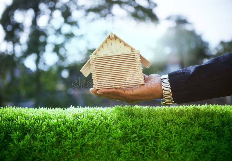 Hand of business man and wood house model on beautiful green grass field use for housing ,residental real estate and land development