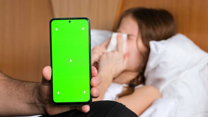 Hand holding phone with green screen mock up, Sick child girl lying on bed on background royalty free stock image
