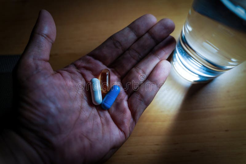 A hand with vitamins minerals and omega 3 supplement capsules next to a glass of water. A male person having health vitamin