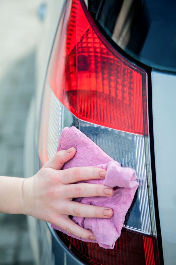 The hand of a young woman holds a pink rag, washes the taillight in a dark grey car. Car wash concept outdoors.