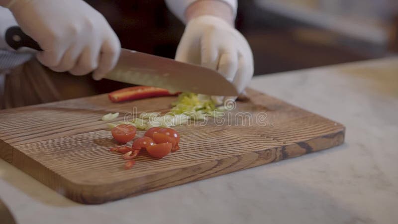 Handchef in den weißen Gummichefhandschuhen schnitt Tomaten und Grüns auf einer hölzernen Küchenbrettnahaufnahme Langsame Bewegun