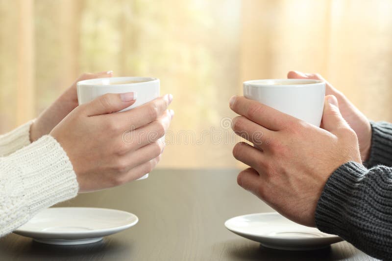 Profile of hands of a couple holding coffee cups over a table in winter in an apartment with a window in the background. Profile of hands of a couple holding coffee cups over a table in winter in an apartment with a window in the background