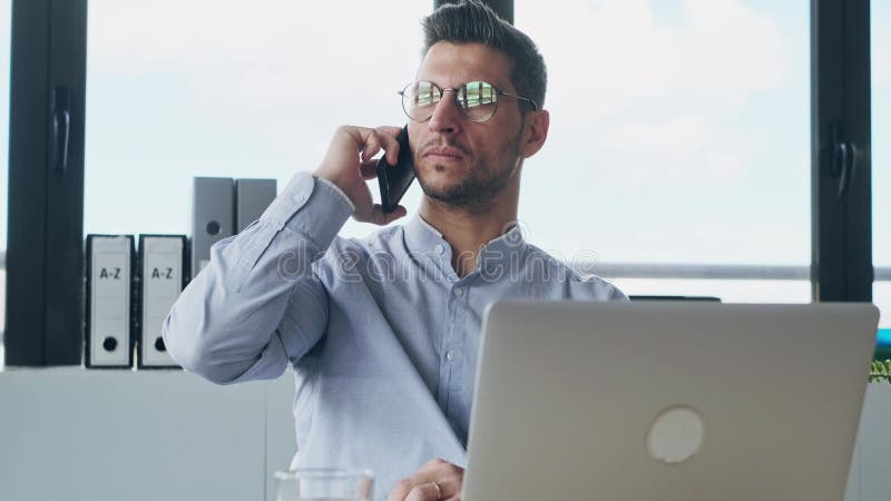 Handige, volwassen ondernemer die praat met smartphone terwijl hij met computer werkt in een modern startkantoor