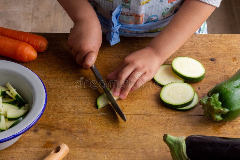 Hands of a child, with an apron, cutting zucchini, helping to prepare the dinner, at a table full of vegetables stock images Hands of a child, with an apron, cutting zucchini, helping to prepare the dinner, at a table full of vegetables stock images