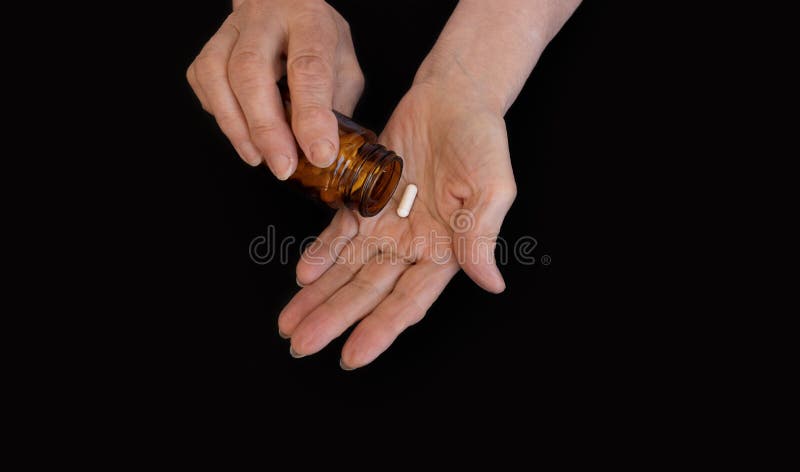 The hands of an elderly woman pour a pill from a jar into her palm. Black background, copy paste for your text. The concept of medicine, diseases of the elderly, prevention, taking vitamins. The hands of an elderly woman pour a pill from a jar into her palm. Black background, copy paste for your text. The concept of medicine, diseases of the elderly, prevention, taking vitamins