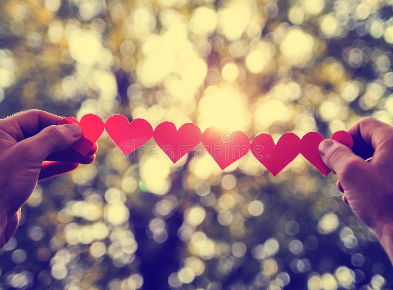 Hands holding a string of paper hearts up to the sun during sunset toned with a retro vintage instagram filter effect