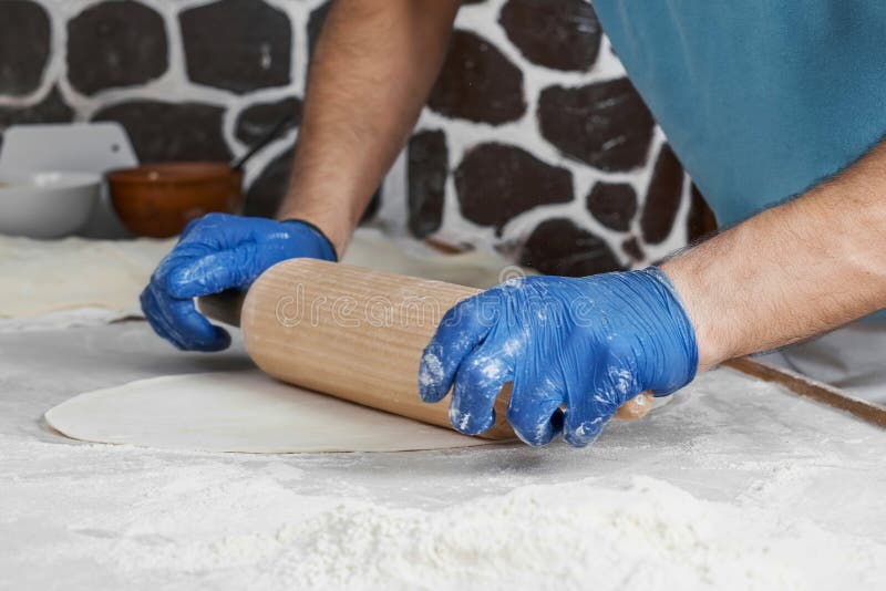 The hands of a male chef in blue gloves roll out the dough on the table. Making traditional Turkish buns, tombik or pide. Close-up