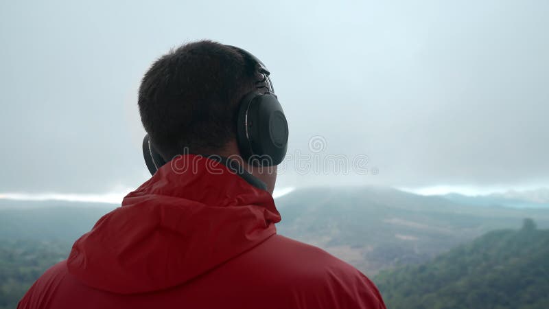 Handsome adult man listening to music through large headphones in mountains on a stormy day looking at valley and misty