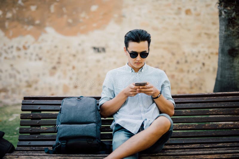 Handsome asian man typing message on his smart phone sitting on stock image Handsome asian man typing message on his smart phone sitting on stock image