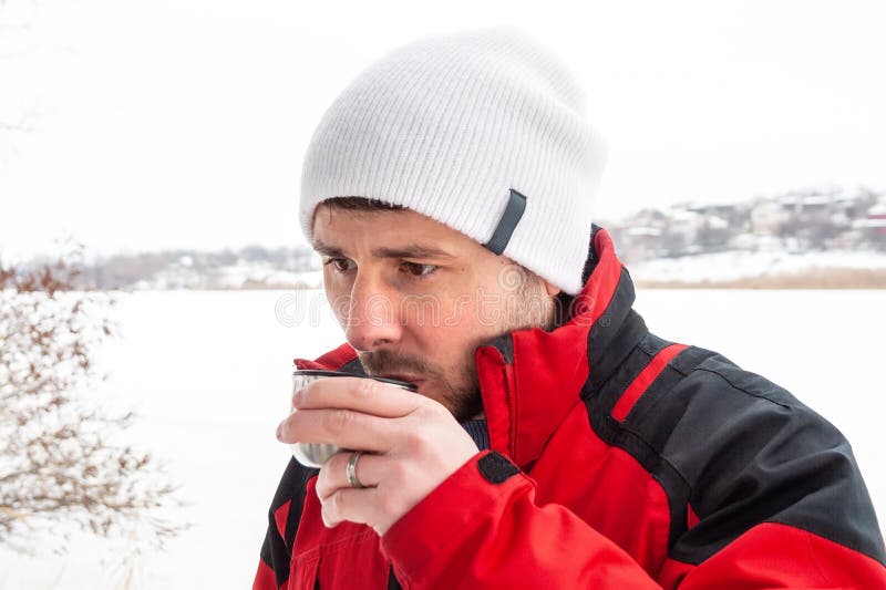 Handsome bearded man in a red and black jacket and white cap drinking hot tea from a metal cup against a frozen river stock image Handsome bearded man in a red and black jacket and white cap drinking hot tea from a metal cup against a frozen river stock image