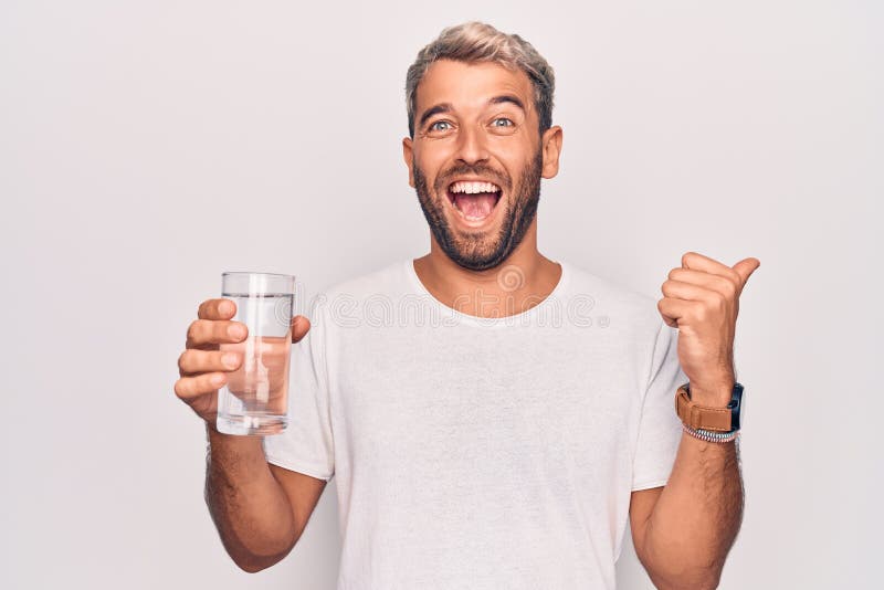 Handsome blond man with beard drinking glass of water to refreshment over white background pointing thumb up to the side smiling