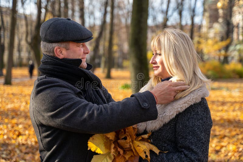 Handsome brunette middle-aged men carefully adjusts his hair. Happy blonde mature woman. Loving couple 45-50 years old are walking in the park, looking at each other. Handsome brunette middle-aged men carefully adjusts his hair. Happy blonde mature woman. Loving couple 45-50 years old are walking in the park, looking at each other