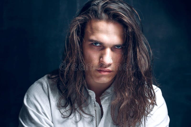 Handsome calm man. Strong boy on an isolated dark background in the studio. Handsome calm man. Portrait of a young muscular guy with long hair. Strong boy on an
