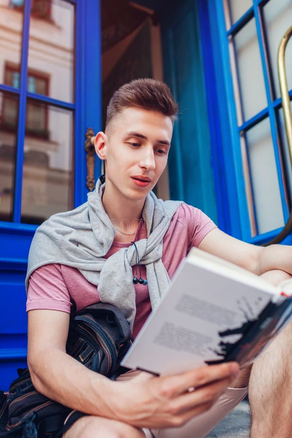 Handsome college man reading a book sitting by cafe. Happy guy student with backpack learning outdoors. Education stock photo Handsome college man reading a book sitting by cafe. Happy guy student with backpack learning outdoors. Education stock photo