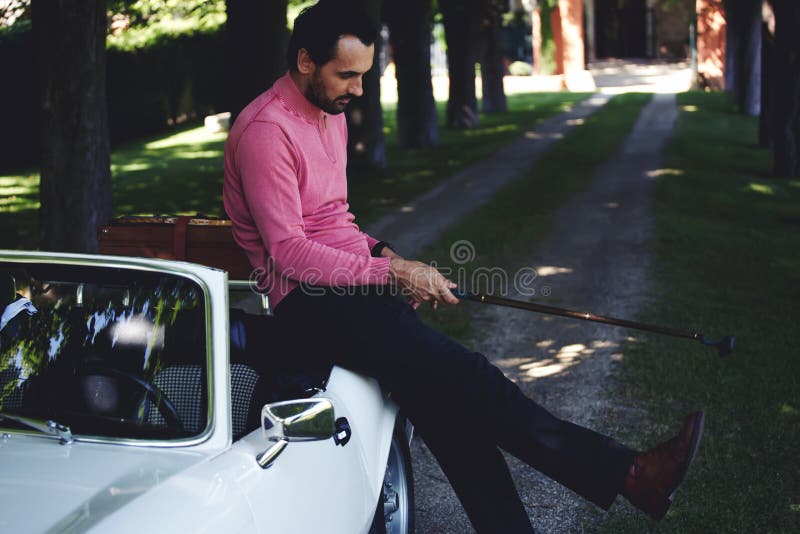 Handsome golf player holding a driver or golf club while getting ready for a day on the course, wealthy man leaning on his convertible luxury car preparing for golf game at recreation time on weekend