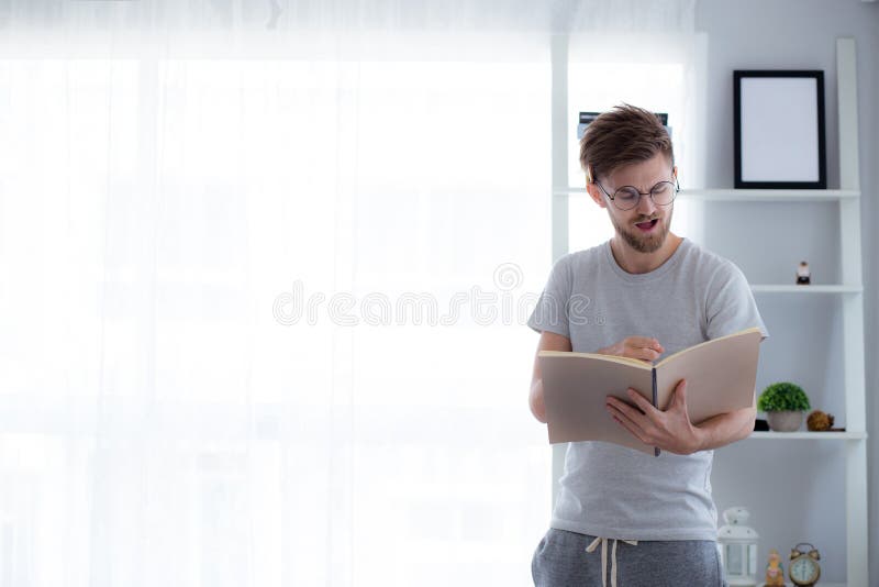 Handsome guy in eyeglasses is reading book preparing exam and thinking with standing at the living room.