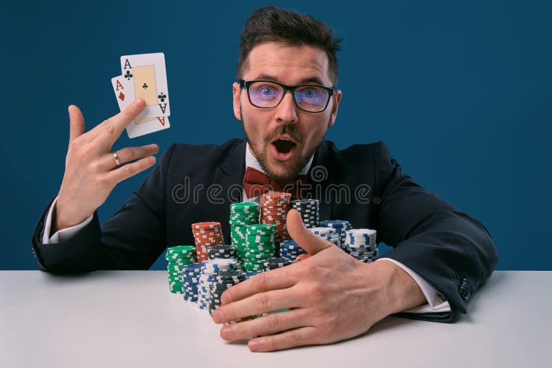 Guy in glasses, black suit sitting at white table with stacks of chips, holding two playing cards, posing on blue stock photography