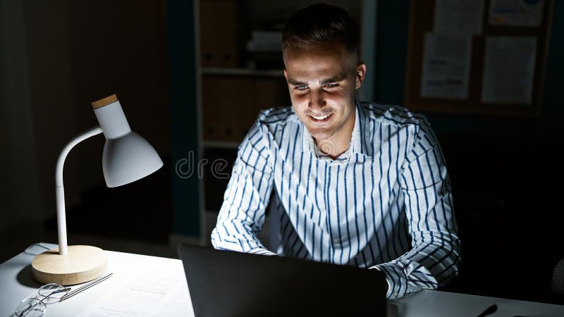 Handsome hispanic man smiling while working on laptop in a well-lit office at night stock photos