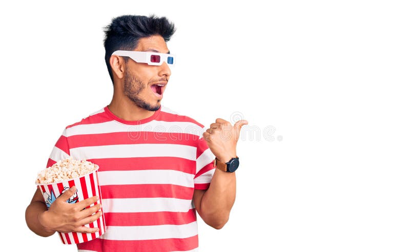 Handsome latin american young man wearing 3d glasses and eating popcorn at the movies pointing thumb up to the side smiling happy