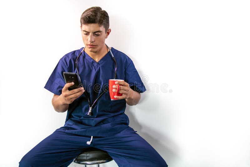 Handsome male doctor in blue scrubs, sitting on stool using phone while drinking from mug stock images Handsome male doctor in blue scrubs, sitting on stool using phone while drinking from mug stock images