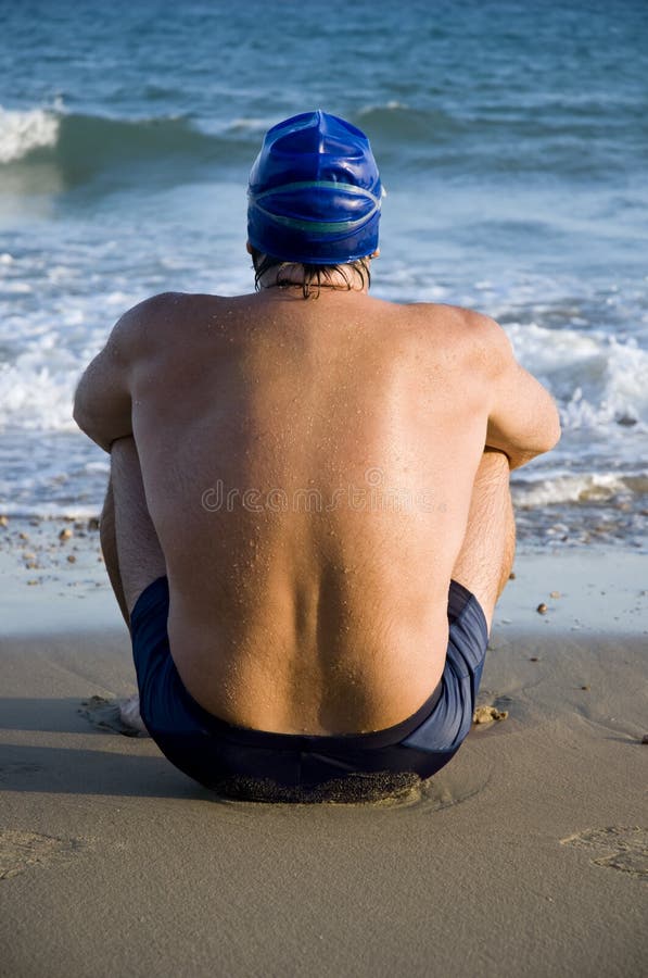 A colour portrait of a handsome macho looking swimmer wearing a blue cap and goggles and looking out to sea while sitting on the beach. A colour portrait of a handsome macho looking swimmer wearing a blue cap and goggles and looking out to sea while sitting on the beach.