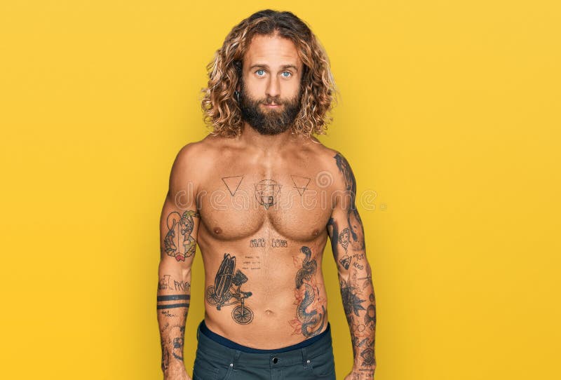 Handsome man with beard and long hair standing shirtless showing tattoos relaxed with serious expression on face