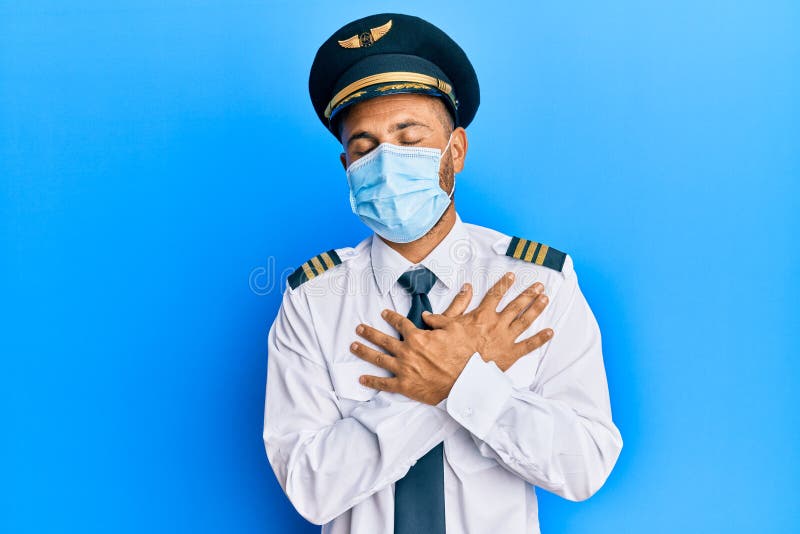 Handsome man with beard wearing airplane pilot uniform wearing safety mask smiling with hands on chest with closed eyes and