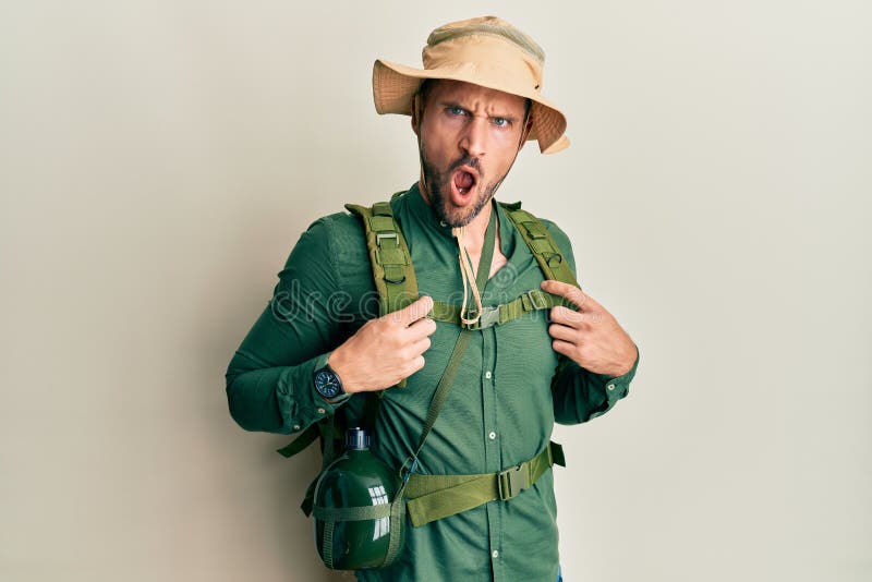 Handsome man with beard wearing explorer hat and backpack in shock face, looking skeptical and sarcastic, surprised with open royalty free stock image