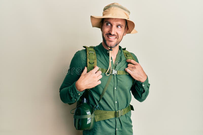 Handsome man with beard wearing explorer hat and backpack smiling looking to the side and staring away thinking stock image