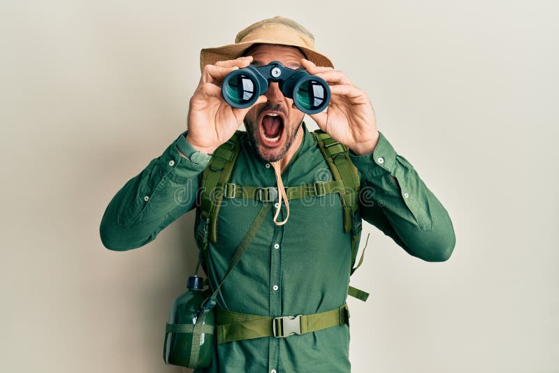 Handsome man with beard wearing explorer hat looking through binoculars celebrating crazy and amazed for success with open eyes stock photography