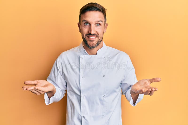 Handsome man with beard wearing professional cook uniform clueless and confused expression with arms and hands raised stock photo