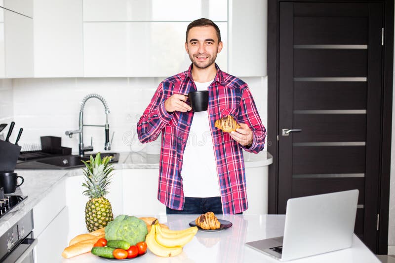 Handsome man with cup of tea or coffee and laptop computer sitting at kitchen royalty free stock images