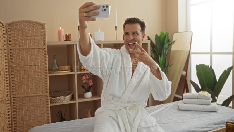 Handsome man making victory gesture while taking selfie in a spa wellness center, wearing a white robe, indoors with relaxing ambiance, suggesting a wellness and soothing experience. Pampering experience stock images, royalty-free photos and pictures