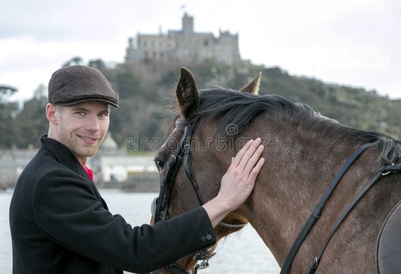 Handsome man, Male Horse Rider pets his horse on beach, wearing traditional flat cap, white trousers, red polo shirt