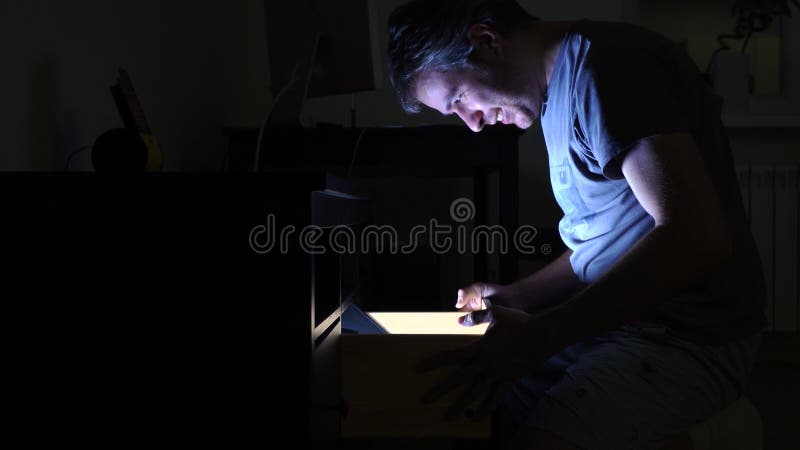 Handsome man opening illuminated drawer in dark room, profile view. Search, find, quest, and surprise concepts. 4K video