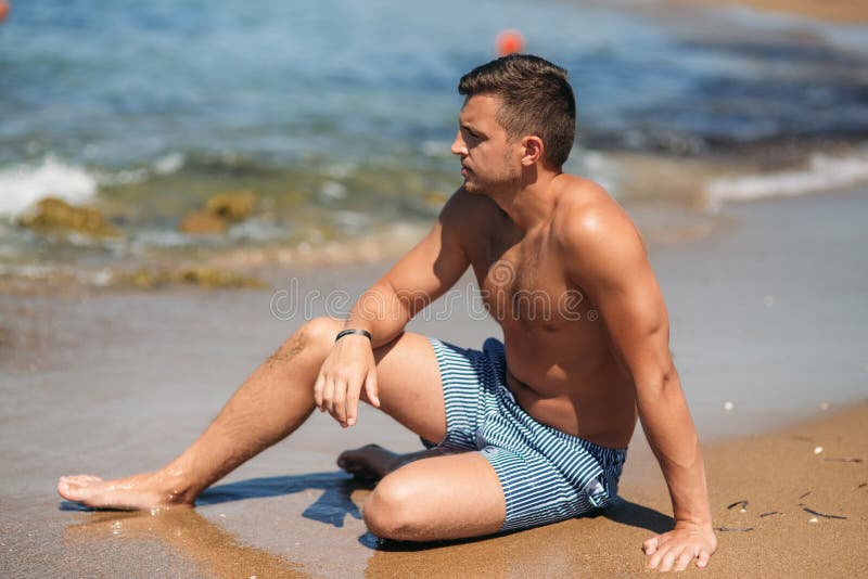 Handsome man in shorts sitting on the sand beach near rocks on the background of sea