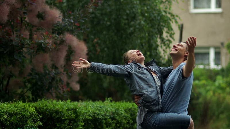 Handsome Man Swings His Girlfriend on the Arms During a Rainy Day in the City Park. Young Couple Enjoying the Rain